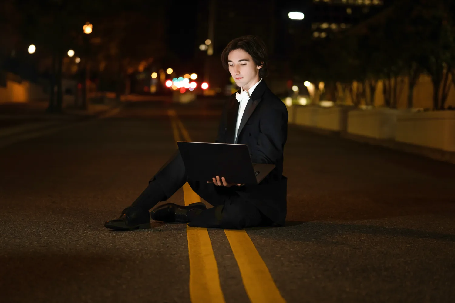 Cade in a tuxedo sitting in an empty city road at night, working on a laptop
